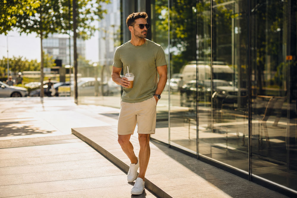 Mężczyzna w letniej stylizacji Recea: szałwiowy t-shirt premium, beżowe spodenki chino i białe sneakersy, spacerujący po mieście w słoneczny majowy dzień.